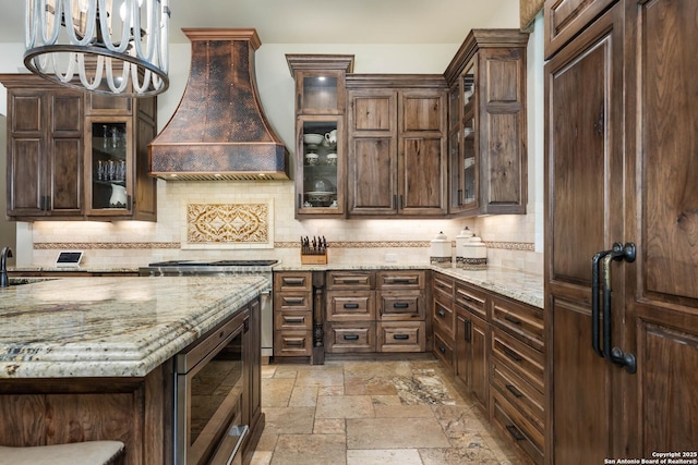 kitchen featuring glass insert cabinets, custom exhaust hood, light stone counters, and stone tile flooring