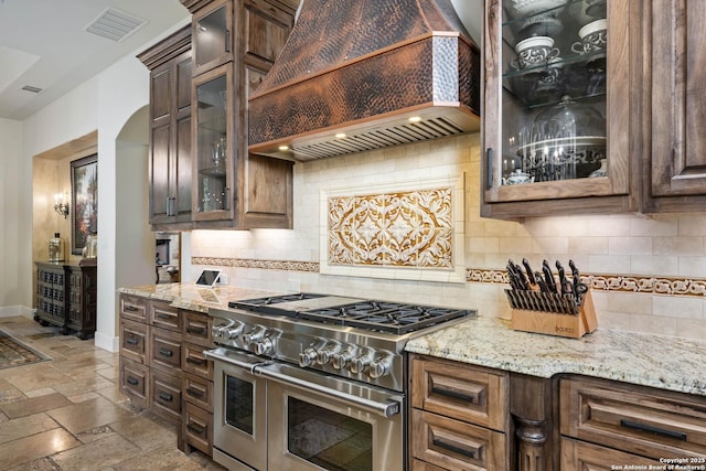 kitchen with stone tile floors, glass insert cabinets, light stone countertops, double oven range, and premium range hood