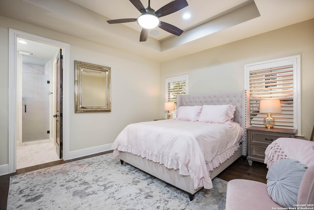 bedroom with a tray ceiling, a ceiling fan, dark wood-type flooring, ensuite bath, and baseboards