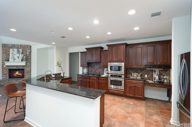 kitchen featuring sink, a brick fireplace, decorative backsplash, a center island with sink, and appliances with stainless steel finishes