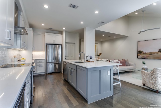 kitchen with wall chimney exhaust hood, stainless steel appliances, sink, a center island with sink, and dark hardwood / wood-style floors