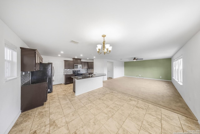 kitchen with tasteful backsplash, ceiling fan with notable chandelier, dark brown cabinetry, stainless steel appliances, and a kitchen island with sink