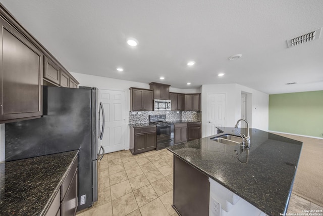 kitchen featuring appliances with stainless steel finishes, dark stone counters, dark brown cabinets, a kitchen island with sink, and sink