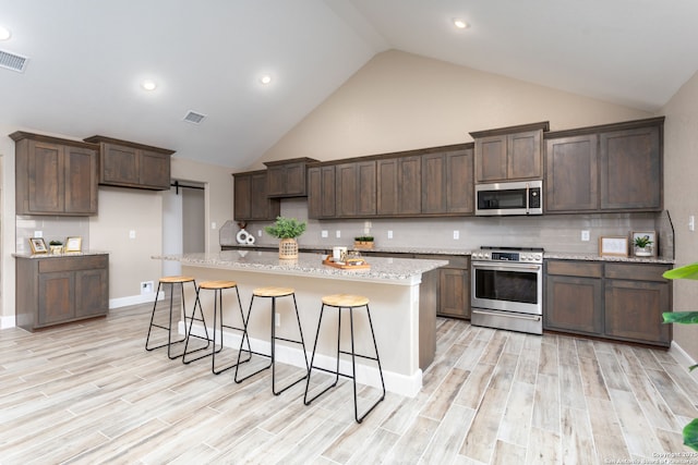 kitchen featuring decorative backsplash, appliances with stainless steel finishes, light wood-type flooring, a kitchen island with sink, and high vaulted ceiling