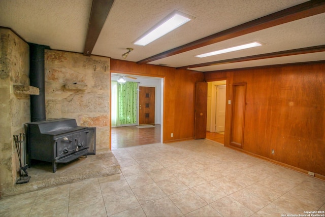 unfurnished living room featuring a textured ceiling, a wood stove, light tile patterned floors, wood walls, and beam ceiling