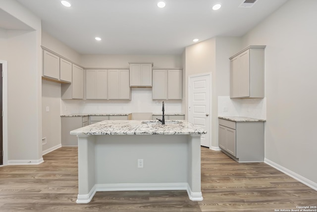 kitchen featuring gray cabinets, light stone counters, a center island with sink, and dark wood-type flooring