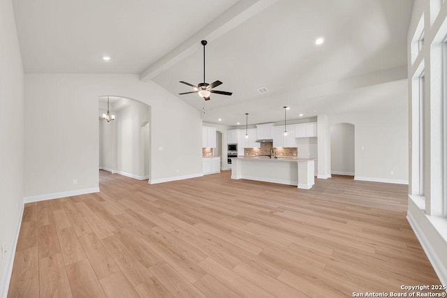 unfurnished living room featuring arched walkways, vaulted ceiling with beams, light wood-style flooring, baseboards, and ceiling fan with notable chandelier