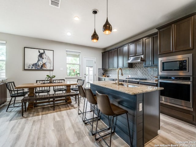 kitchen featuring light stone countertops, appliances with stainless steel finishes, dark brown cabinetry, and a kitchen island with sink