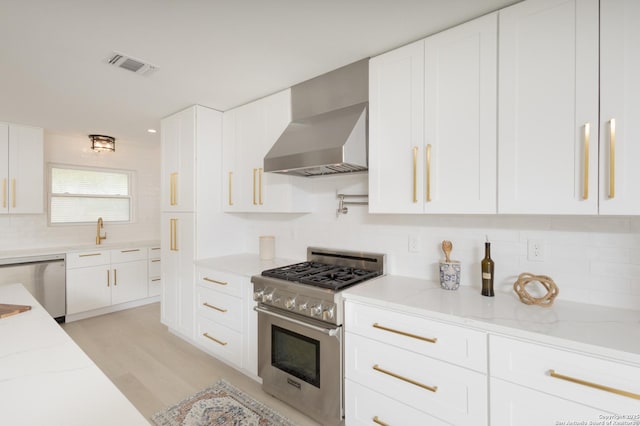 kitchen with light stone countertops, stainless steel appliances, wall chimney range hood, light hardwood / wood-style floors, and white cabinetry