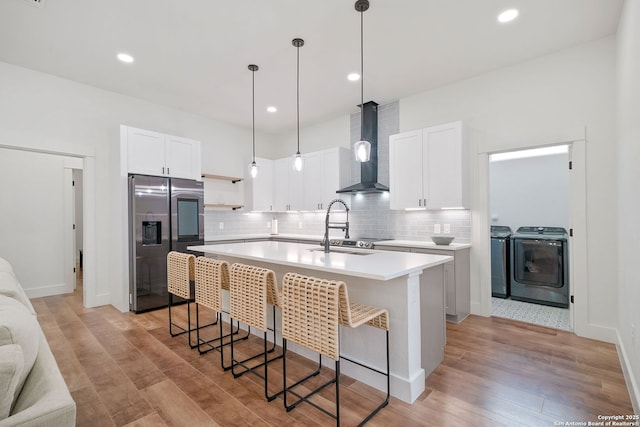 kitchen with wall chimney exhaust hood, stainless steel fridge, separate washer and dryer, an island with sink, and white cabinetry