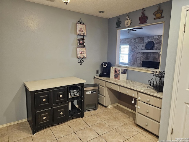 home office featuring ceiling fan, a stone fireplace, a textured ceiling, light tile patterned floors, and built in desk