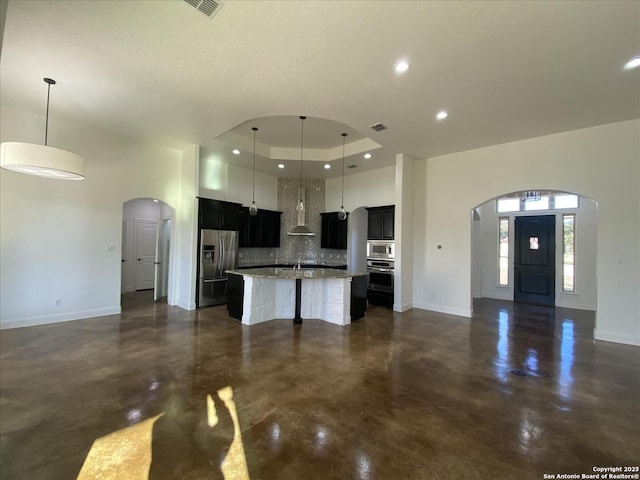 kitchen with stainless steel appliances, decorative light fixtures, a raised ceiling, and a kitchen island with sink