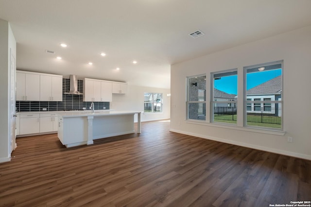 kitchen with wall chimney exhaust hood, dark wood-type flooring, backsplash, an island with sink, and white cabinets