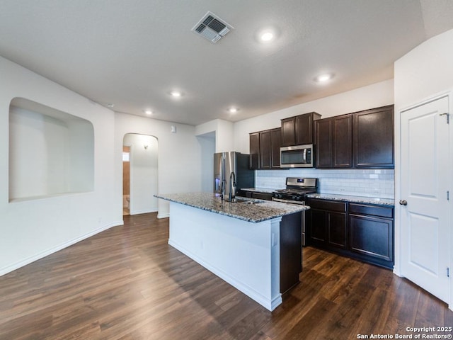 kitchen featuring backsplash, dark hardwood / wood-style floors, dark stone countertops, an island with sink, and stainless steel appliances