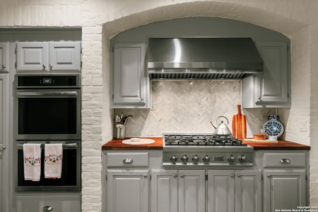 kitchen featuring backsplash, butcher block counters, wall chimney range hood, and appliances with stainless steel finishes