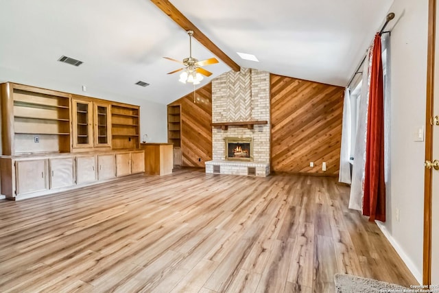 unfurnished living room with wood walls, lofted ceiling with beams, light hardwood / wood-style flooring, a brick fireplace, and ceiling fan