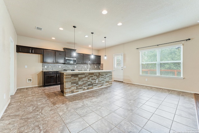 kitchen featuring backsplash, a kitchen island with sink, hanging light fixtures, light tile patterned floors, and light stone countertops