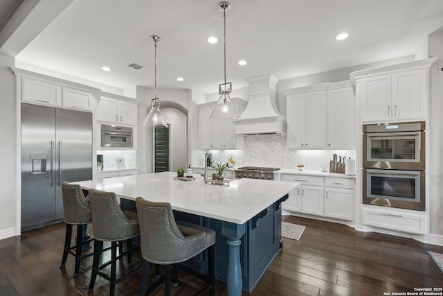 kitchen with stainless steel appliances, white cabinets, custom exhaust hood, hanging light fixtures, and a kitchen island with sink