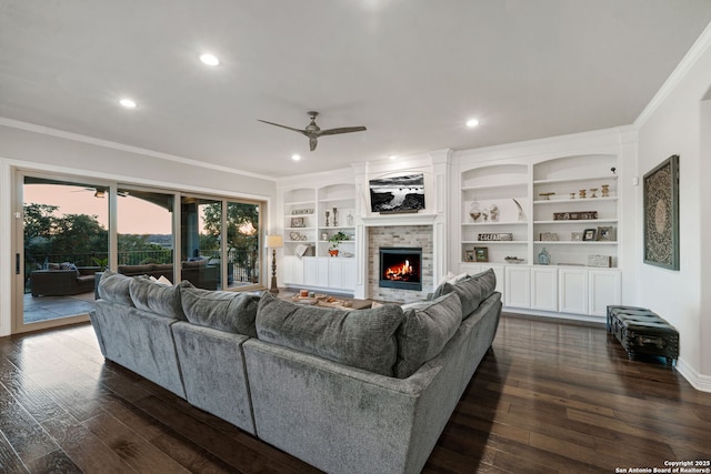 living room featuring a stone fireplace, ceiling fan, built in shelves, crown molding, and dark wood-type flooring