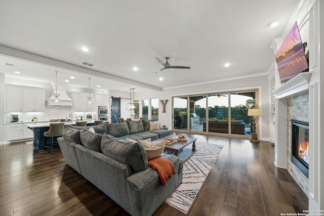 living room with dark wood-type flooring, a brick fireplace, beam ceiling, crown molding, and ceiling fan