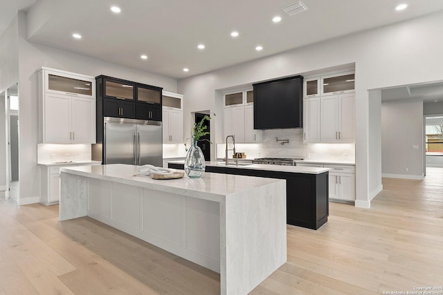 kitchen with built in refrigerator, light stone counters, white cabinetry, and a center island with sink