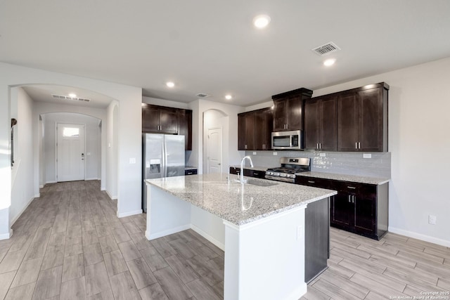 kitchen with light stone counters, stainless steel appliances, an island with sink, light hardwood / wood-style floors, and sink