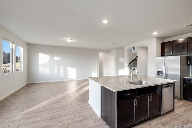 kitchen with sink, light stone counters, an island with sink, dark brown cabinets, and appliances with stainless steel finishes