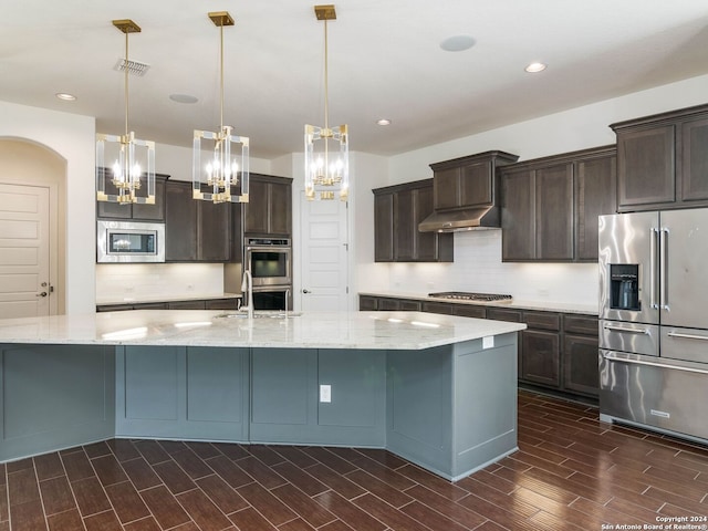 kitchen with stainless steel appliances, a large island with sink, light stone countertops, dark brown cabinets, and hanging light fixtures