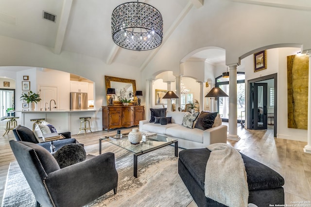 living room with an inviting chandelier, ornate columns, light wood-type flooring, high vaulted ceiling, and beam ceiling