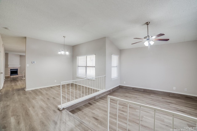 spare room featuring a textured ceiling, vaulted ceiling, light hardwood / wood-style flooring, a brick fireplace, and ceiling fan with notable chandelier