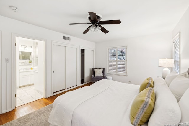 bedroom with light wood-type flooring, two closets, ensuite bath, and ceiling fan