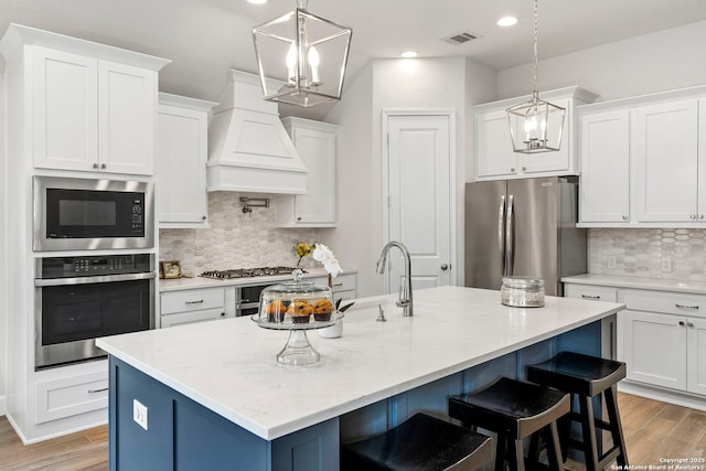 kitchen featuring premium range hood, appliances with stainless steel finishes, a kitchen island with sink, and white cabinetry