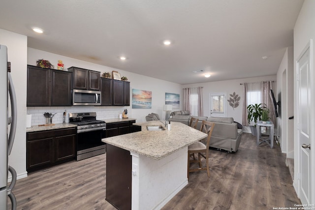 kitchen featuring hardwood / wood-style flooring, stainless steel appliances, a kitchen island with sink, backsplash, and dark brown cabinets