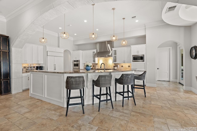 kitchen with built in appliances, decorative light fixtures, white cabinetry, wall chimney exhaust hood, and a spacious island