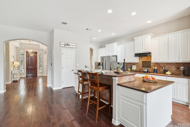 kitchen with dark hardwood / wood-style floors, an island with sink, stainless steel fridge, backsplash, and white cabinetry