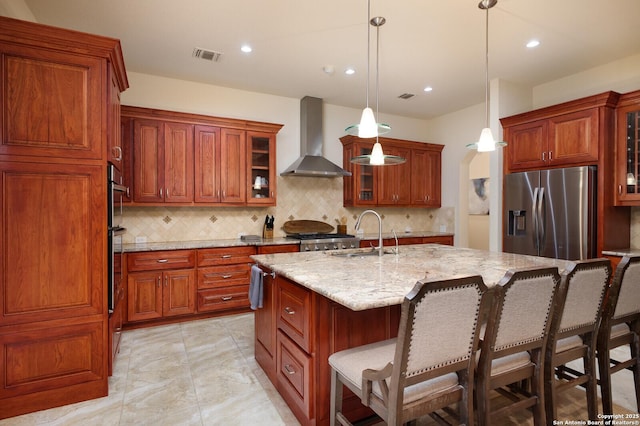 kitchen featuring stainless steel appliances, sink, wall chimney exhaust hood, pendant lighting, and a kitchen island with sink