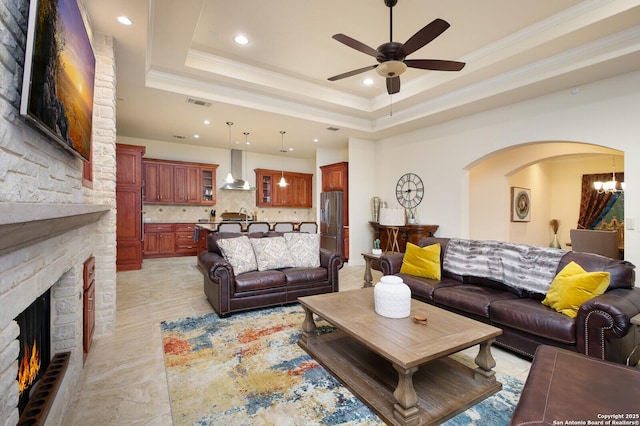 living room with a tray ceiling, ceiling fan with notable chandelier, a fireplace, ornamental molding, and sink