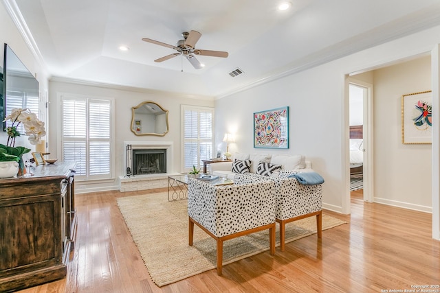 living room featuring a raised ceiling, ceiling fan, ornamental molding, a fireplace, and light hardwood / wood-style flooring