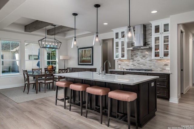 kitchen featuring wall chimney exhaust hood, hanging light fixtures, an island with sink, light hardwood / wood-style floors, and stainless steel gas stovetop