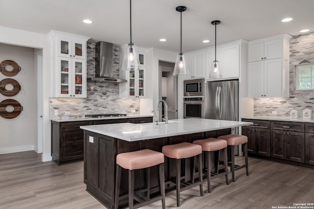 kitchen featuring stainless steel appliances, an island with sink, wall chimney range hood, white cabinets, and sink