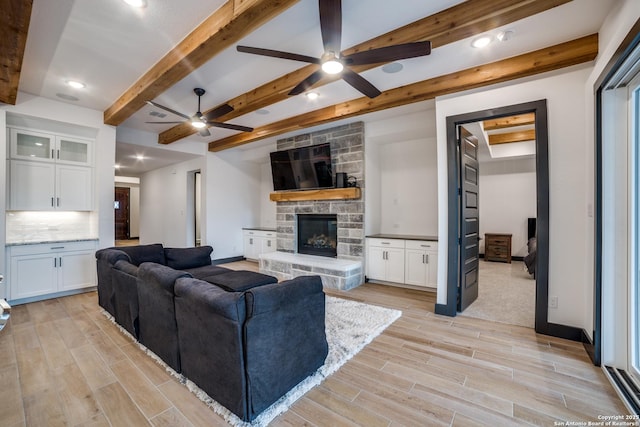 living room featuring beam ceiling, ceiling fan, a stone fireplace, and light hardwood / wood-style floors