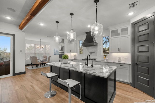 kitchen featuring pendant lighting, premium range hood, white cabinetry, a kitchen island with sink, and light stone countertops