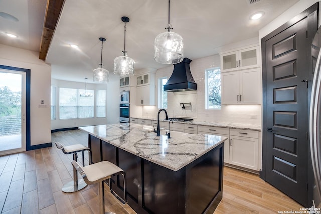 kitchen with white cabinetry, premium range hood, decorative light fixtures, and a kitchen island with sink