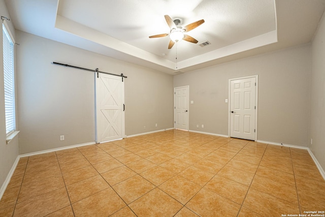 empty room featuring ceiling fan, a barn door, a raised ceiling, and light tile patterned floors