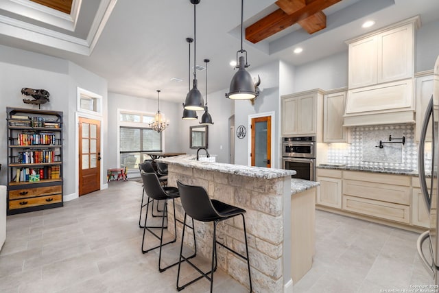 kitchen featuring a kitchen island with sink, cream cabinetry, hanging light fixtures, light stone counters, and an inviting chandelier