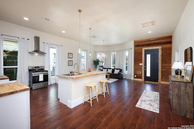 kitchen featuring stainless steel range with gas stovetop, a center island with sink, wall chimney exhaust hood, sink, and butcher block counters