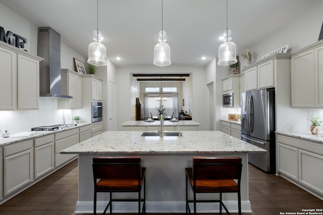 kitchen featuring wall chimney range hood, sink, stainless steel appliances, an island with sink, and decorative light fixtures