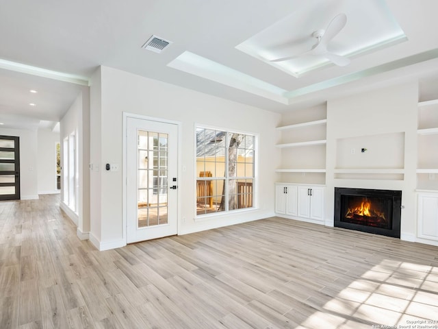 unfurnished living room with a tray ceiling, ceiling fan, built in shelves, and light wood-type flooring