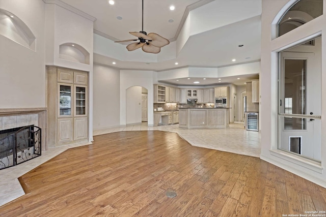 unfurnished living room with a high ceiling, light wood-type flooring, beverage cooler, a tiled fireplace, and ceiling fan