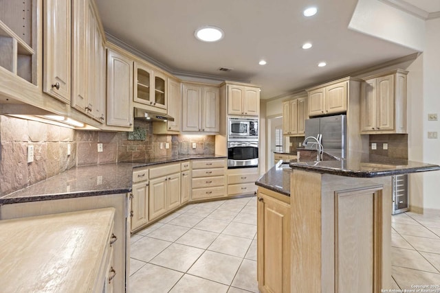 kitchen featuring stainless steel appliances, light brown cabinets, a kitchen island with sink, and dark stone counters